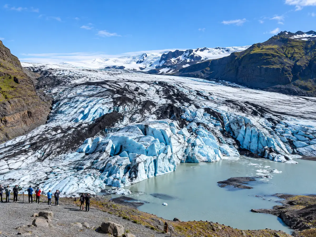 La lengua azul y blanca de Sólheimajökull con bandas de ceniza oscura por encima de una laguna de deshielo, y pequeños visitantes en un mirador